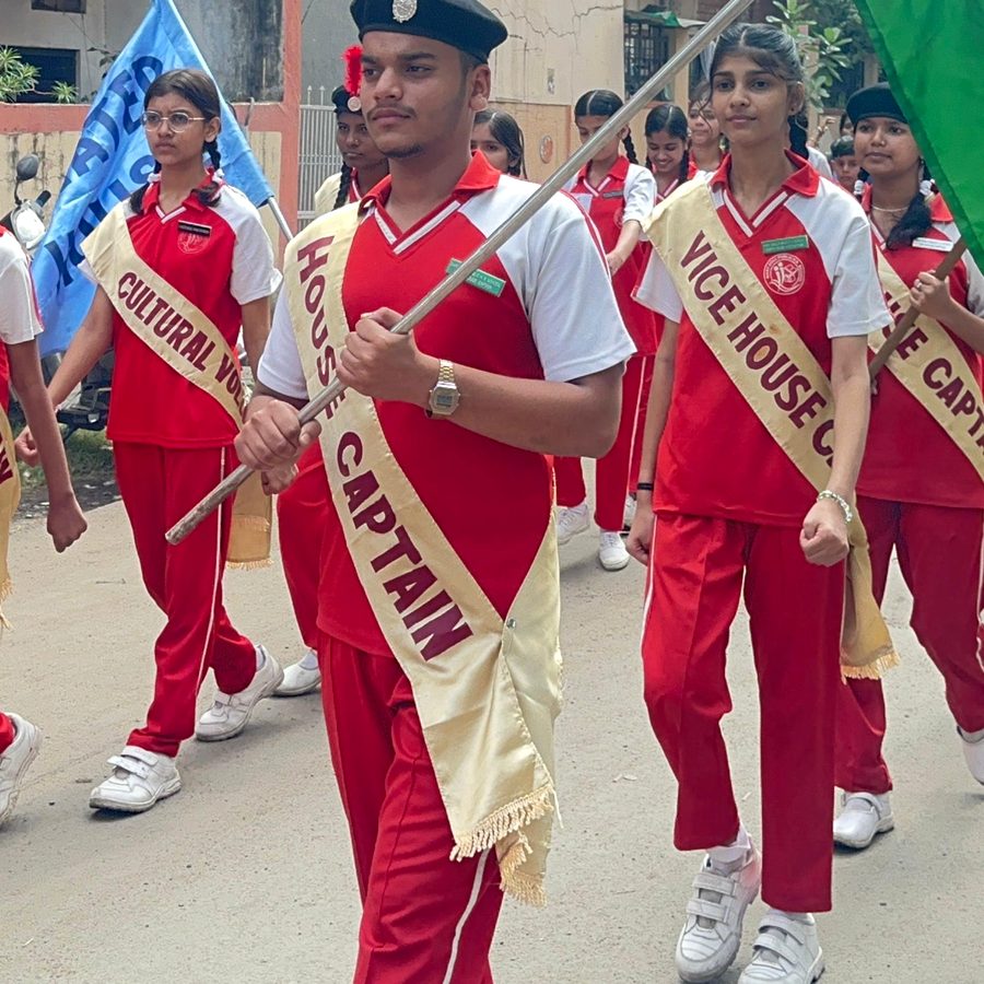 A house captain in a red sash leading the march, holding the house flag